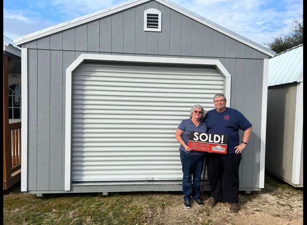 A man and a woman are standing in front of a garage holding a sold sign.