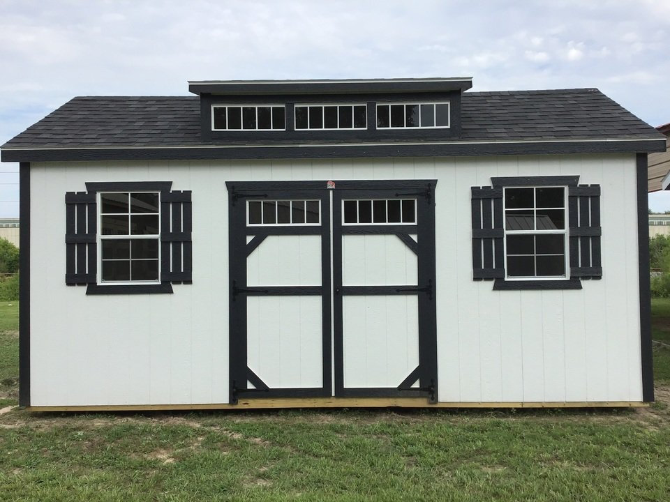 A white shed with black windows and shutters is sitting on top of a lush green field.