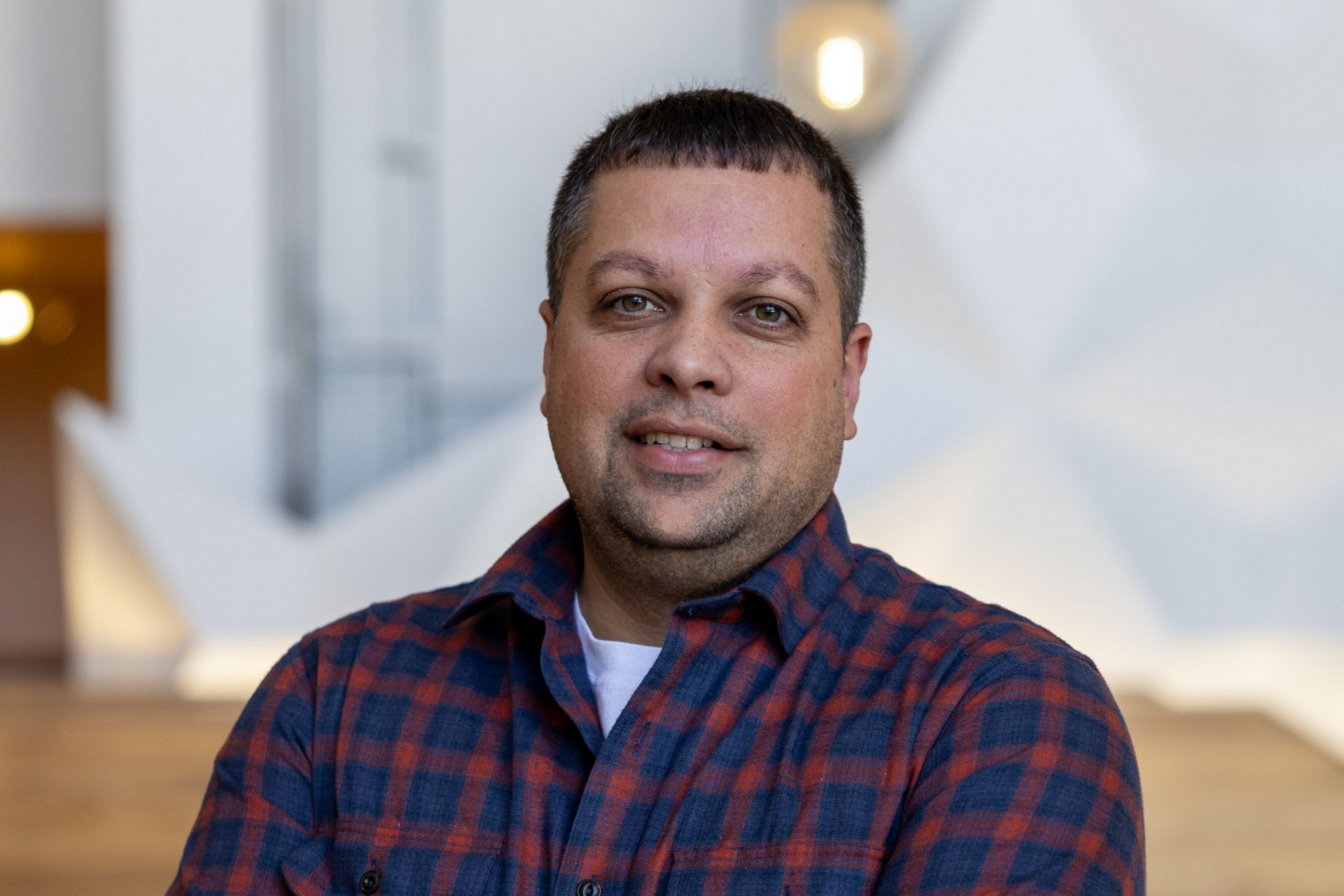 Man in a red and blue plaid shirt seated indoors, facing the camera with a slight smile.