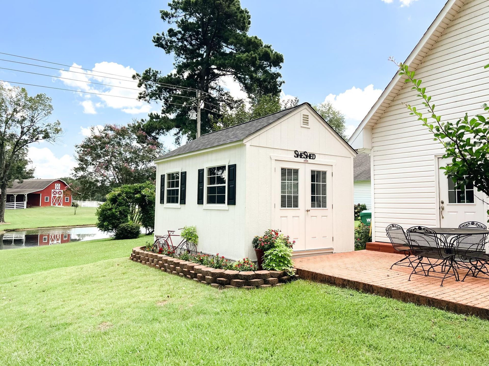 Small white garden shed with black shutters beside a house and patio in a sunny yard
