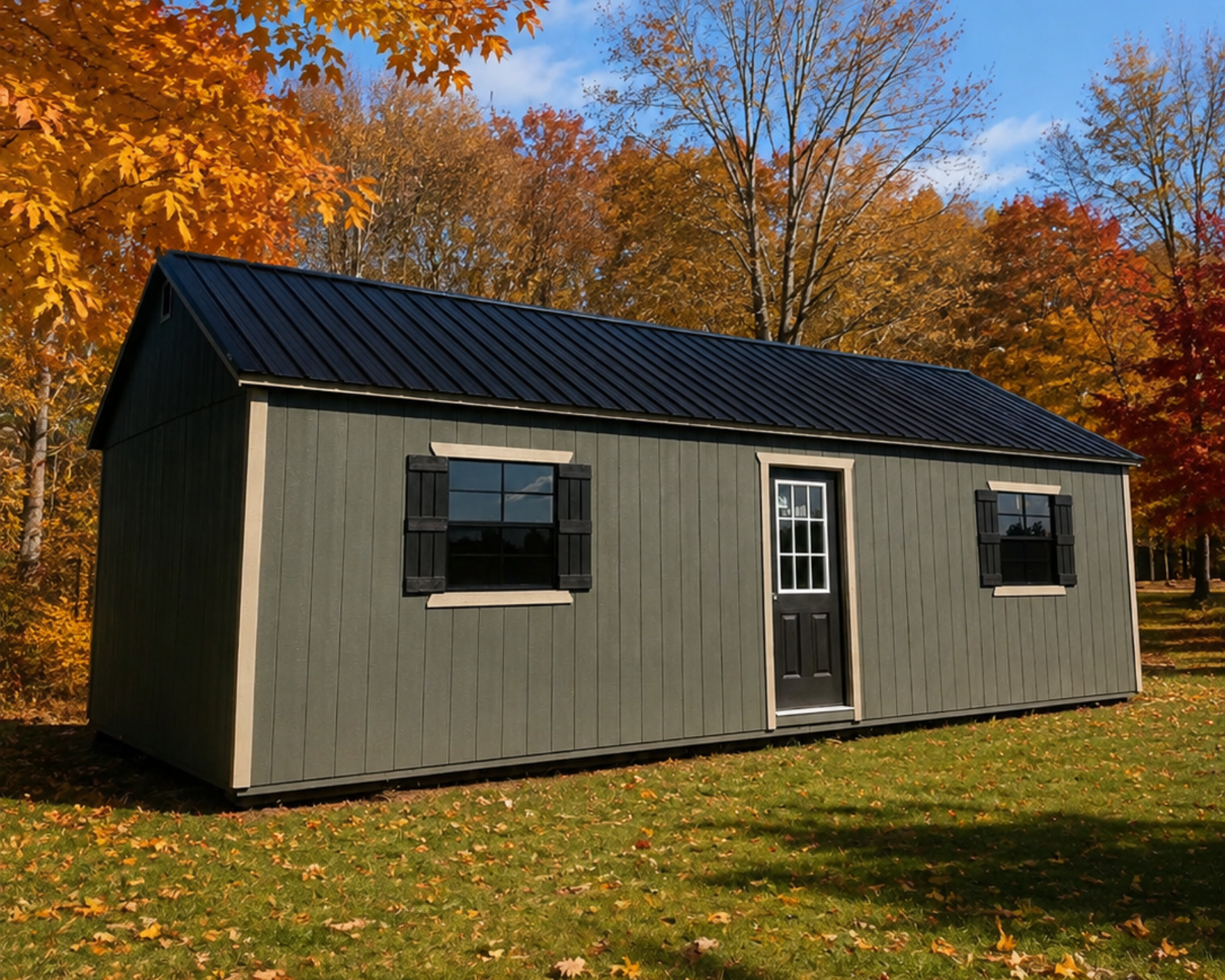Small gray shed with a black roof in a grassy yard, surrounded by autumn trees under a blue sky