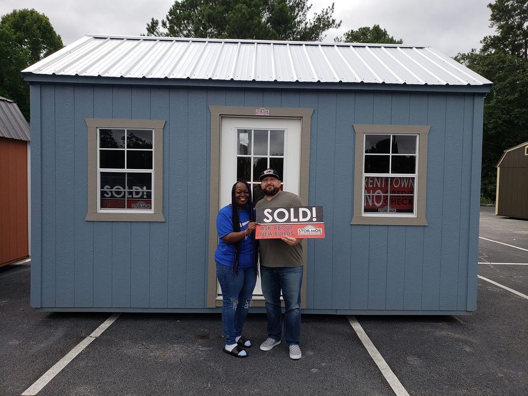 A man and woman are standing in front of a blue shed holding a sold sign.