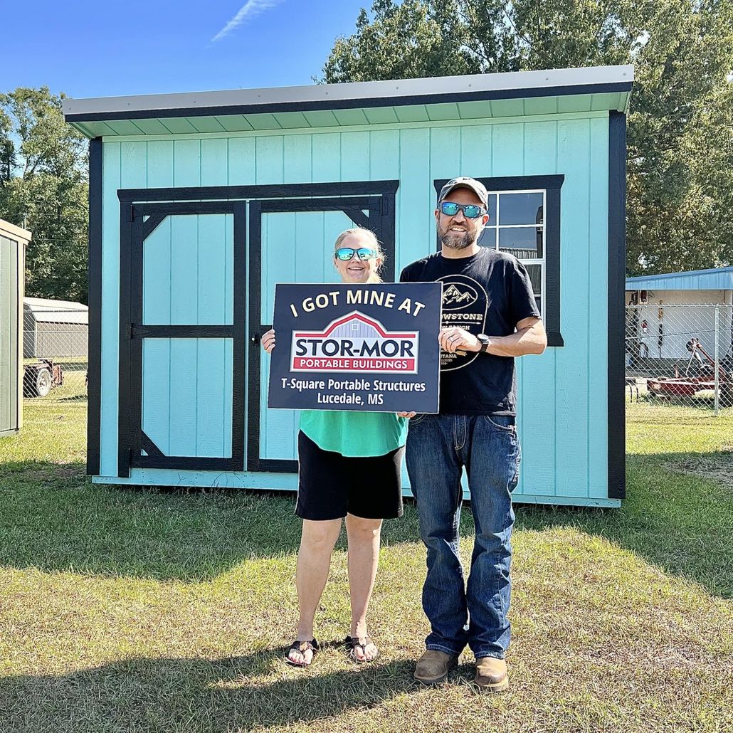 A man and a woman are holding a sold sign in front of a blue shed.
