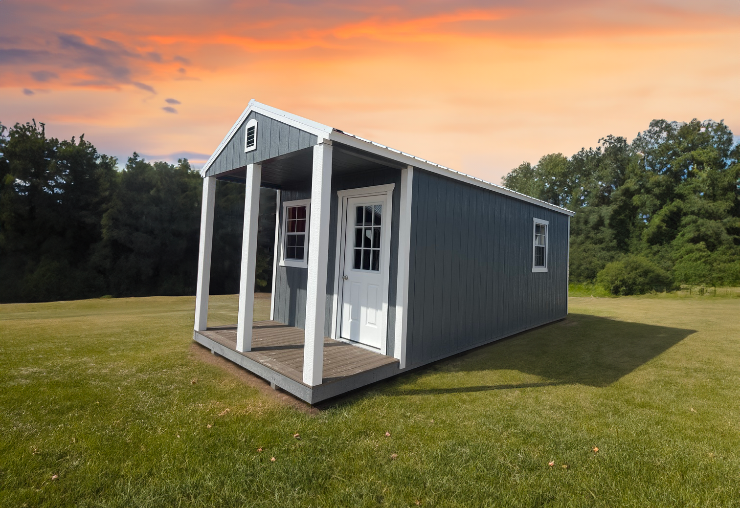 A cabin shed with a porch is sitting in the middle of a grassy field at sunset.