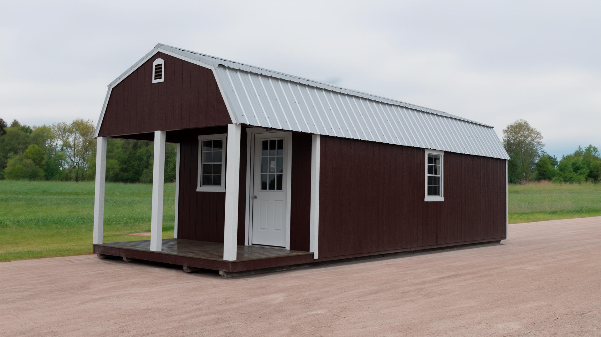 A brown lofted barn with a porch and windows is sitting on top of a dirt road.