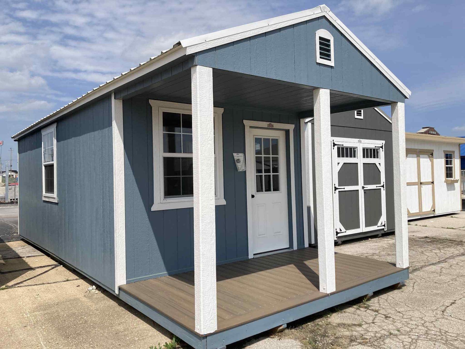 Small blue Cabin with white trim and porch on a concrete lot under a clear sky with other shed options