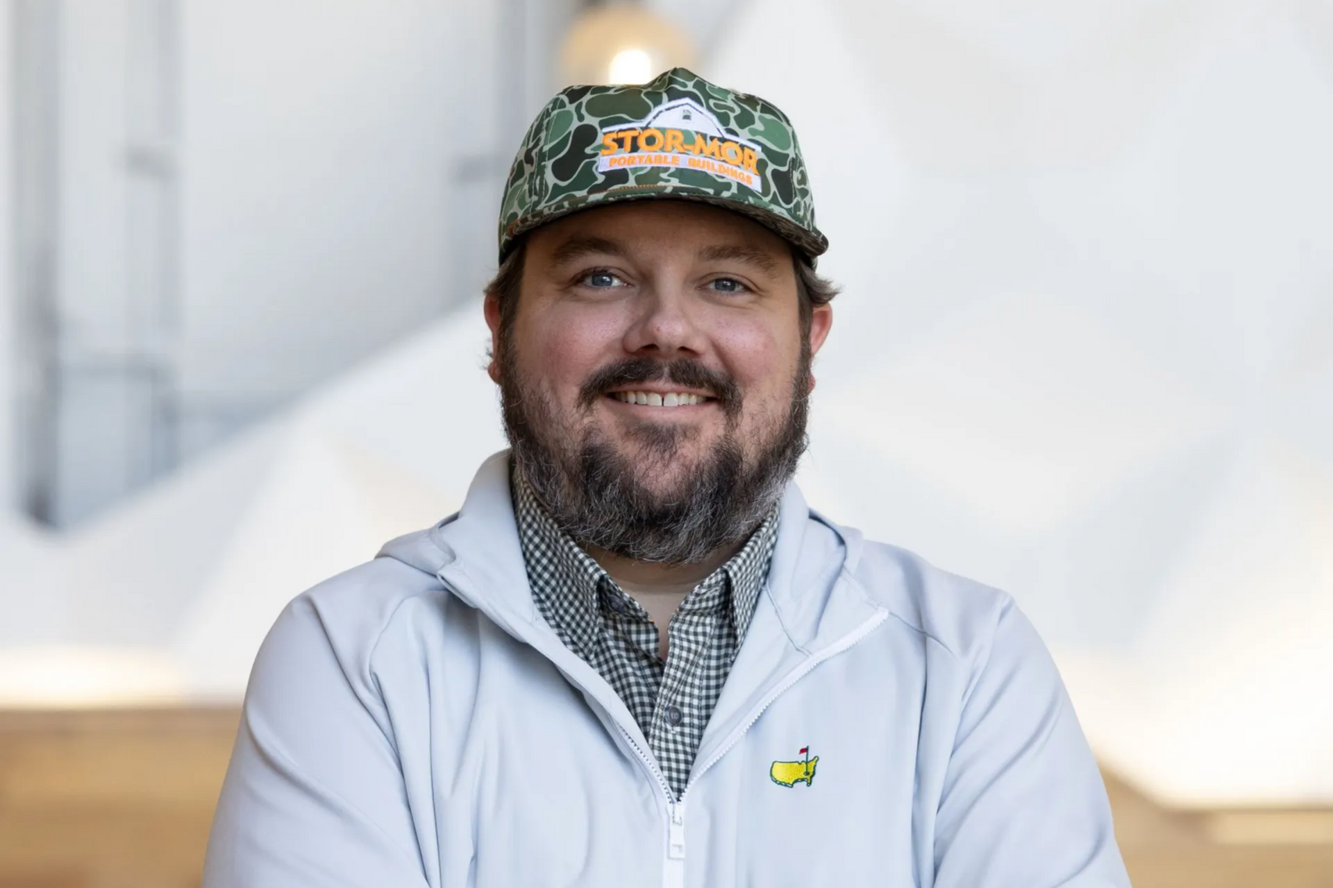 Smiling man in a white jacket and green cap standing indoors with arms crossed