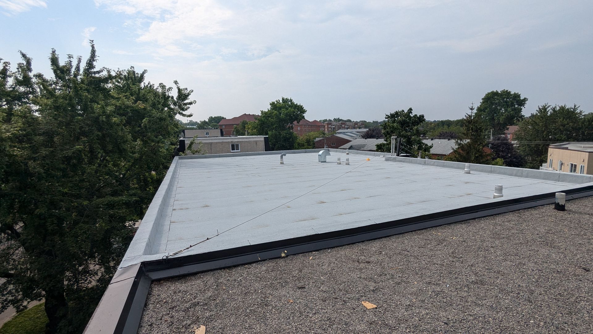 Toit plat recouvert de gravier avec une section en membrane blanche, arbres et bâtiments en arrière-plan
