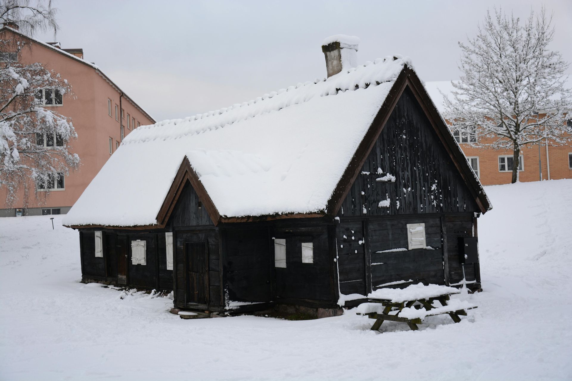Petite cabane noire au toit enneigé dans une cour enneigée, avec des immeubles d'appartements en arrière-plan.