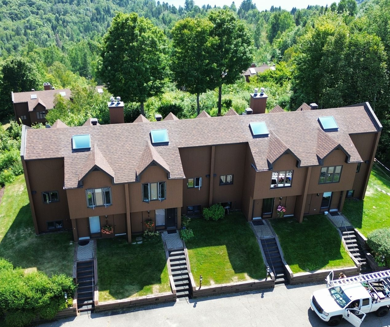 Vue aérienne de maisons de ville brunes aux toits pointus, avec des arbres verts à l'arrière et des voitures garées devant.