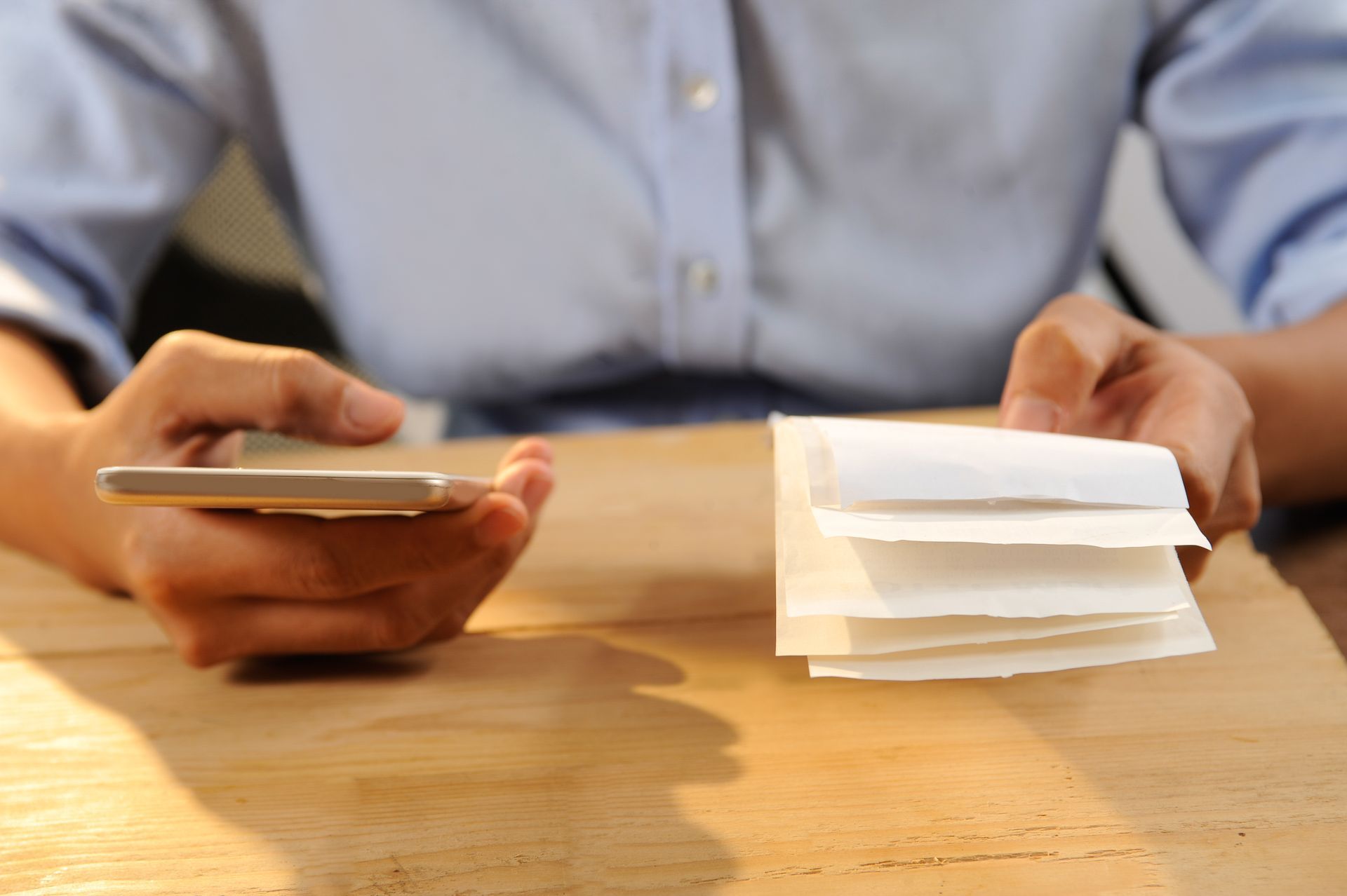 Person holding a smartphone beside a stack of paper documents on a wooden table
