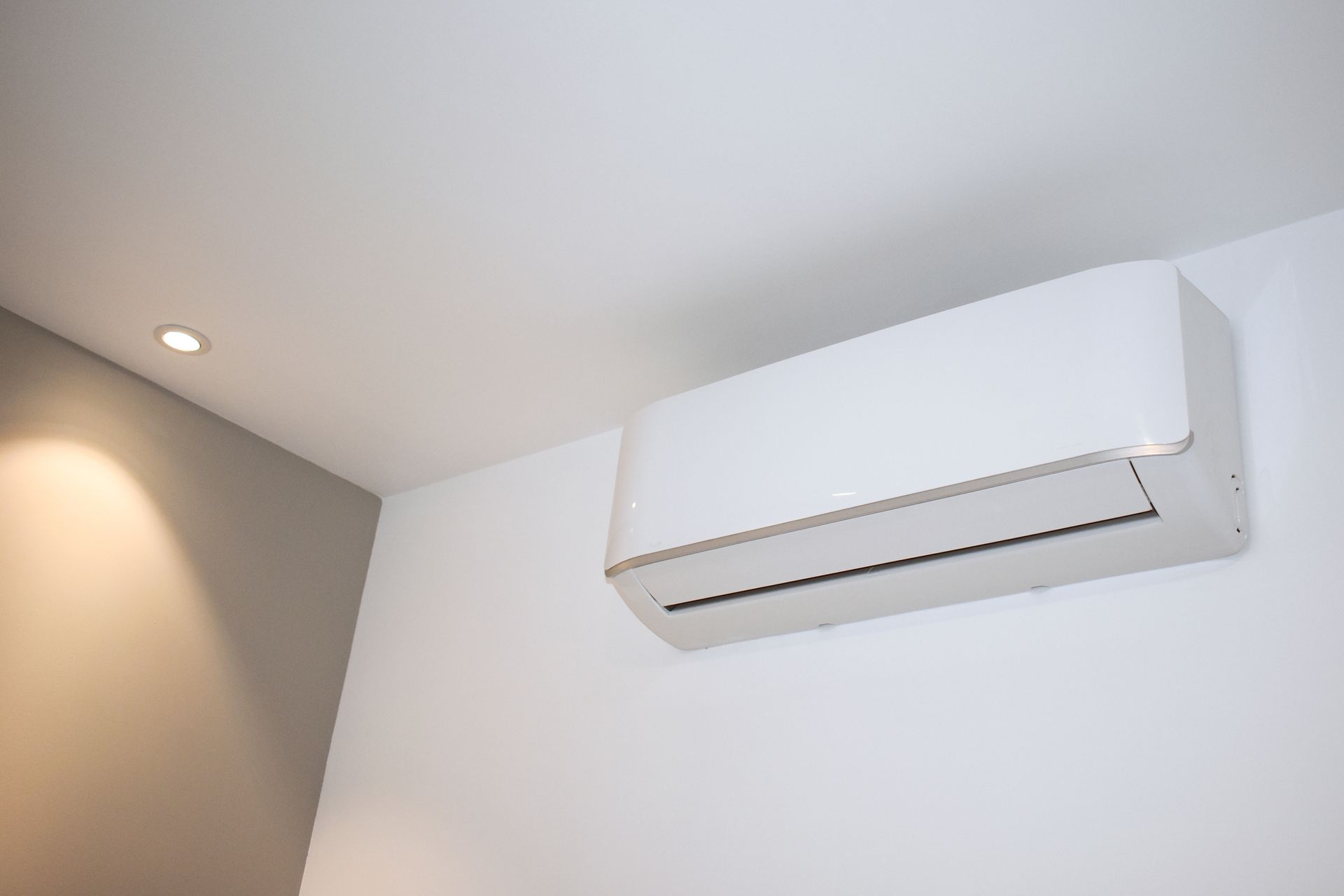 Wall-mounted white air conditioner on a white interior wall near the ceiling, with a recessed light nearby