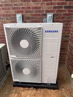 A samsung air conditioner is sitting in front of a brick wall.