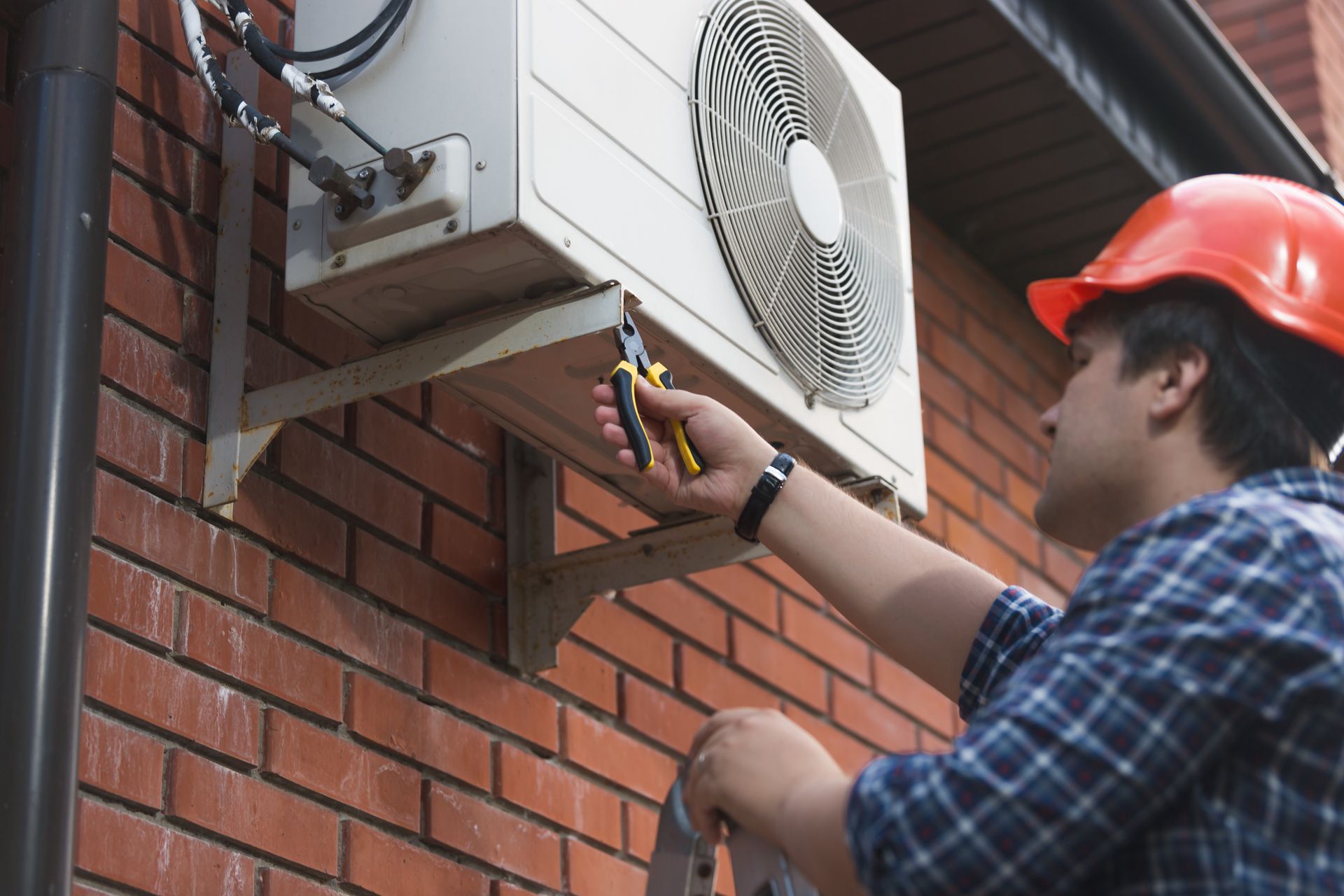 A man wearing a hard hat is fixing an air conditioner on a brick wall.