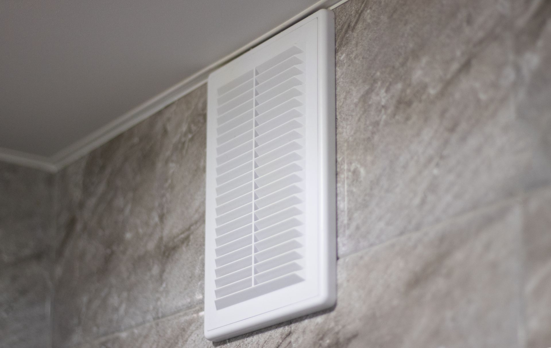 A white air vent is hanging on a tiled wall.