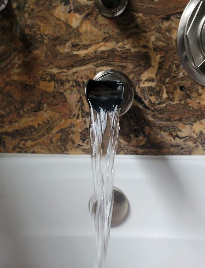 Water flows from a modern, wide-mouthed bathtub faucet mounted on a brown, marbled tile wall.