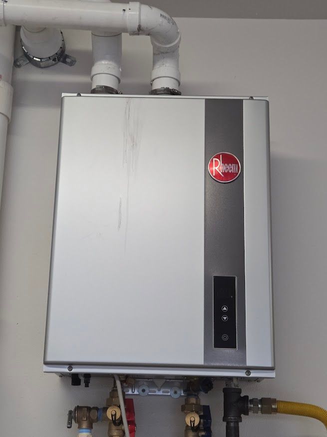 A silver Rheem tankless water heater mounted on a white wall with white PVC venting pipes above and plumbing connections.