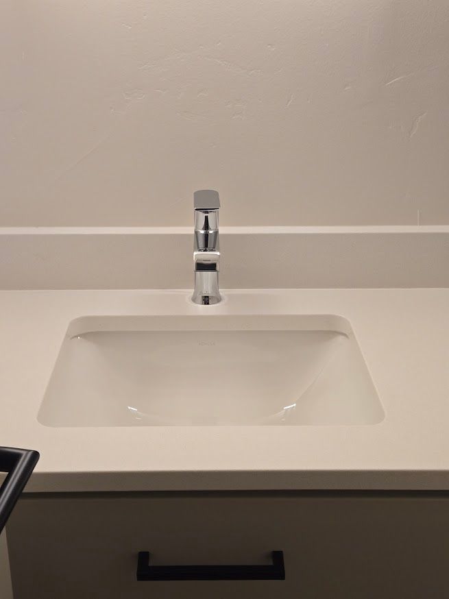 A white bathroom sink with a chrome faucet set in a light-colored countertop above a vanity with a black handle.