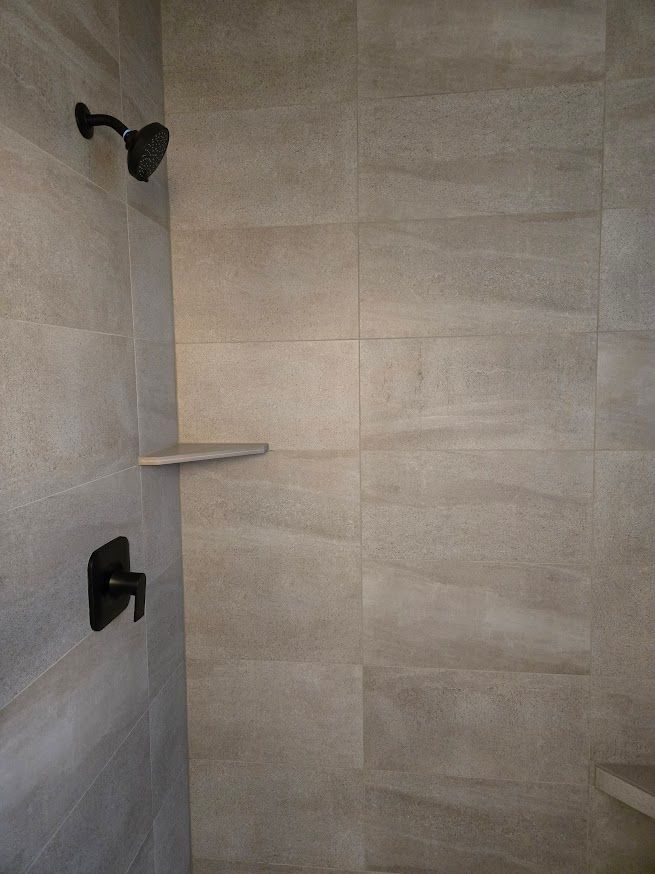 A shower stall with neutral-toned, textured rectangular wall tiles, featuring a matte black shower head and faucet.