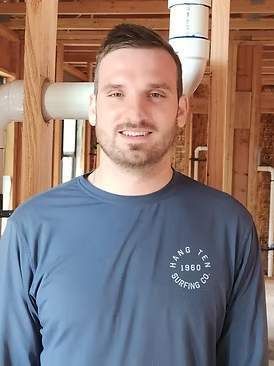 A person smiling while wearing a long-sleeved blue shirt, standing inside an unfinished room with wooden wall studs.