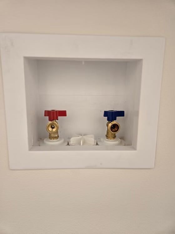 A white laundry supply box inset in a wall, containing brass water valves with red and blue handles.