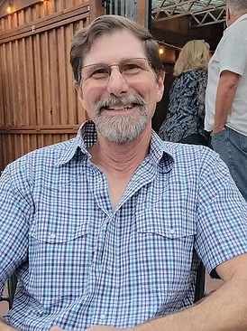 A man with a short beard and glasses wearing a blue and white checkered short-sleeved shirt smiles at an outdoor event.