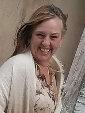 A smiling person with long hair wearing a patterned top and a light-colored cardigan, outdoors.