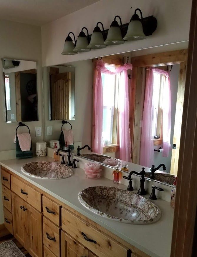A dual-sink bathroom vanity with wood cabinets, stone-style basin sinks, dark fixtures, and pink curtains reflected in mirror.