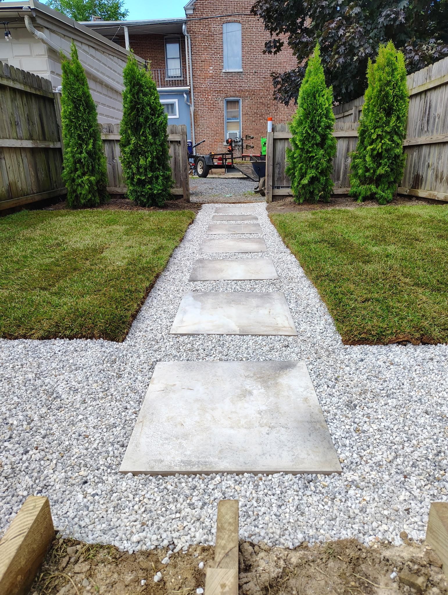 Gravel backyard path with stepping stones leading to a brick house, bordered by grass and tall shrubs.