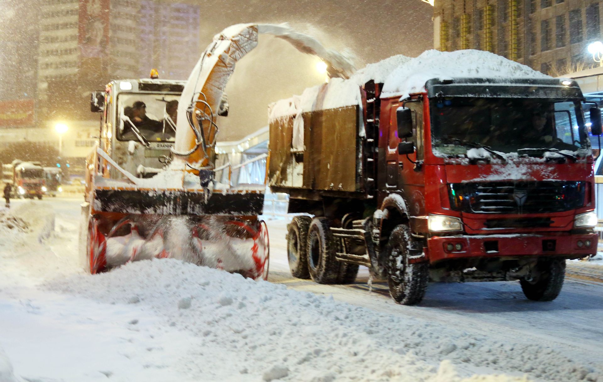 Professional Landscaping Snow blower loading snow into a red dump truck during a heavy snowfall in a city.