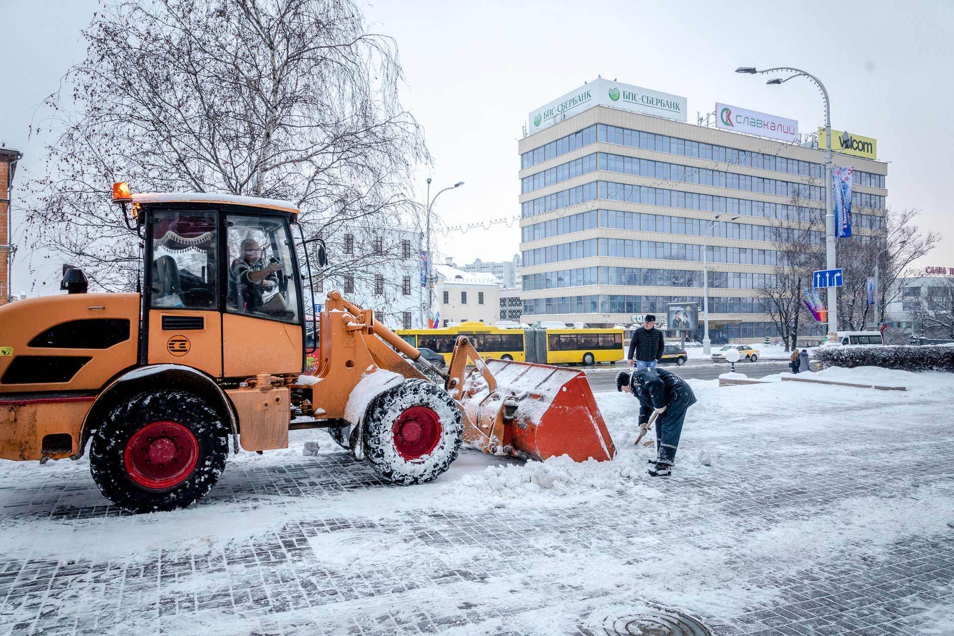 A snowplow and worker clear snow from a street in front of a building during a snowfall.