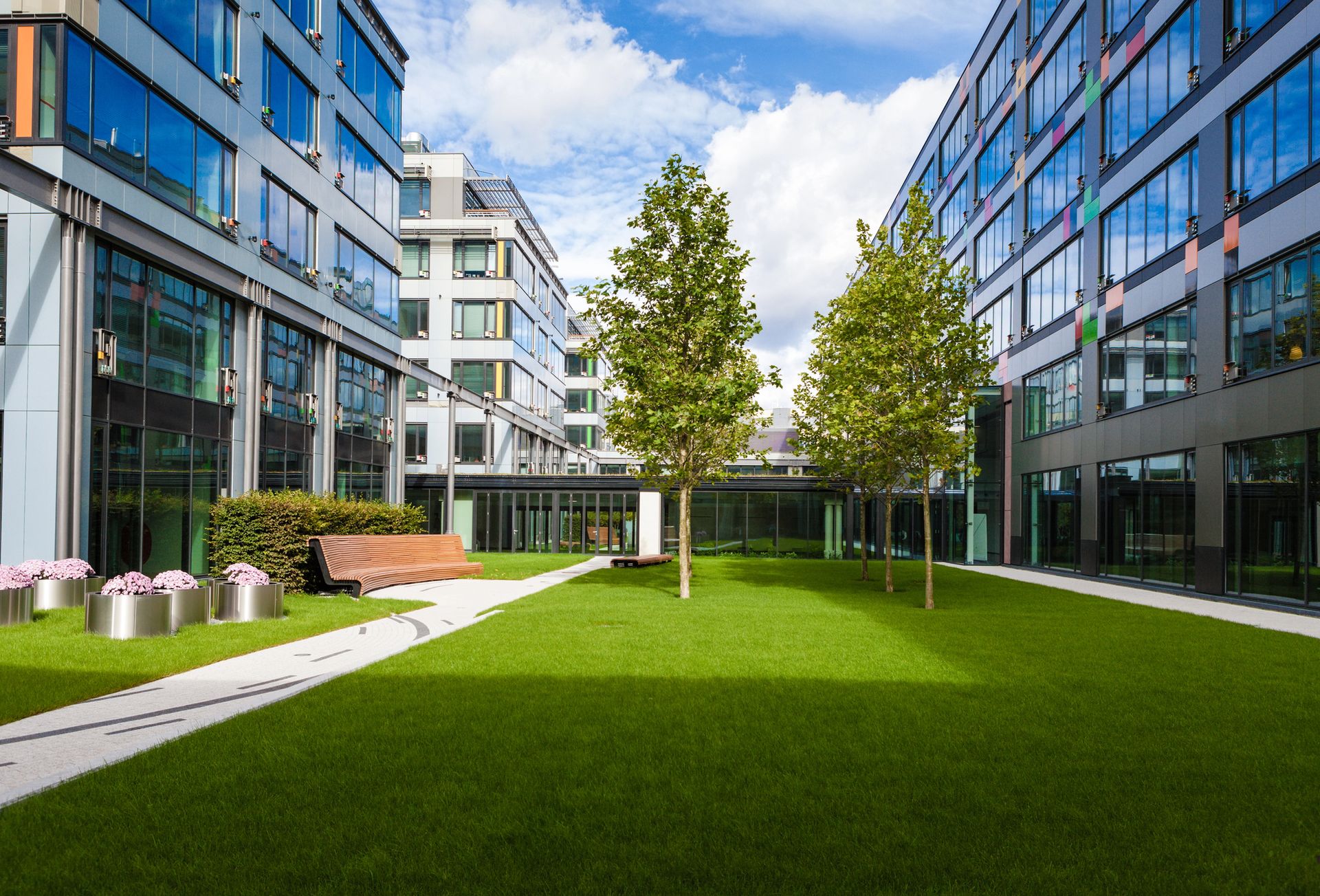Courtyard with green lawn, trees, benches, and modern buildings under a cloudy sky.