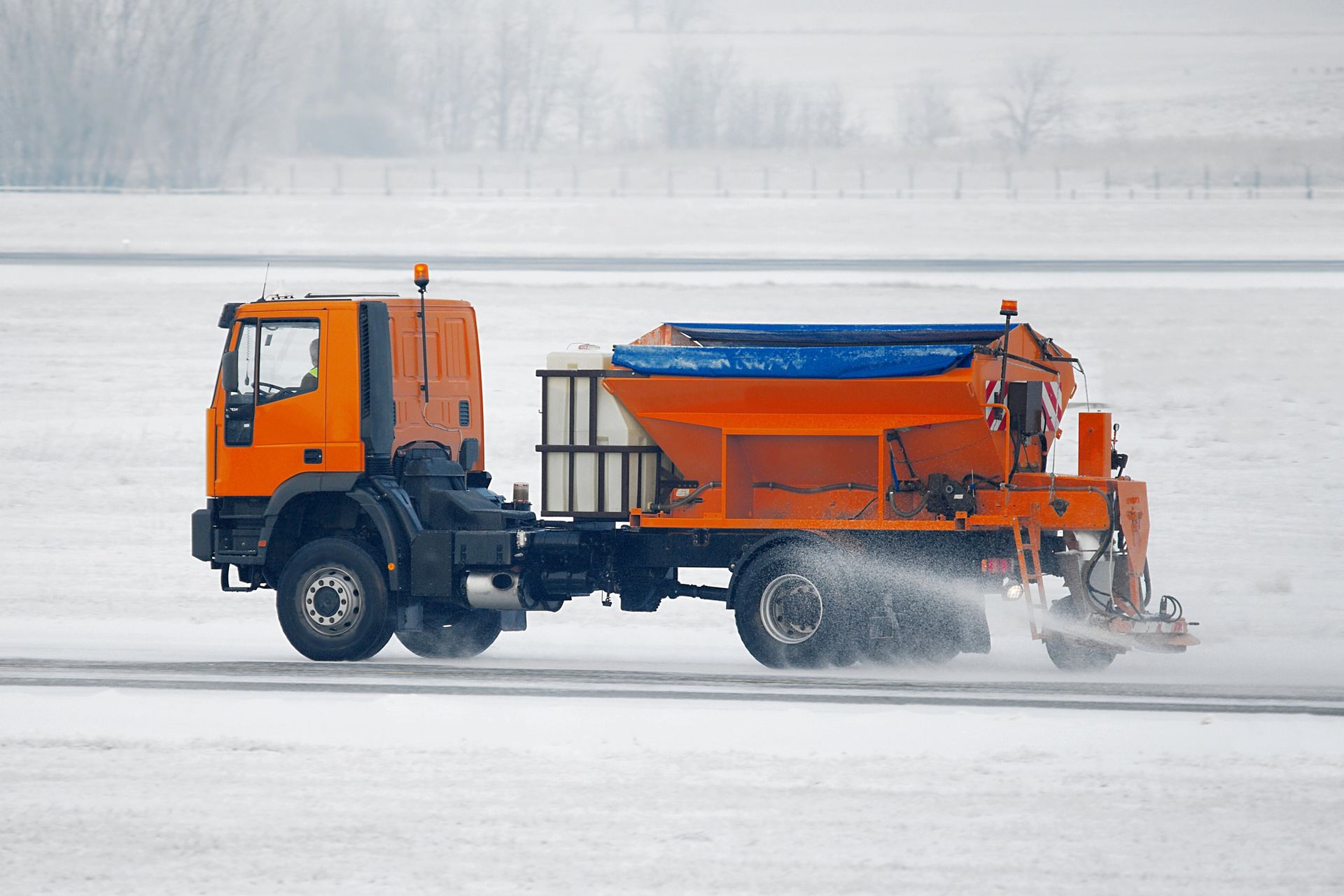 Property Clean Ups Orange snowplow truck spraying de-icing liquid on a snow-covered road.