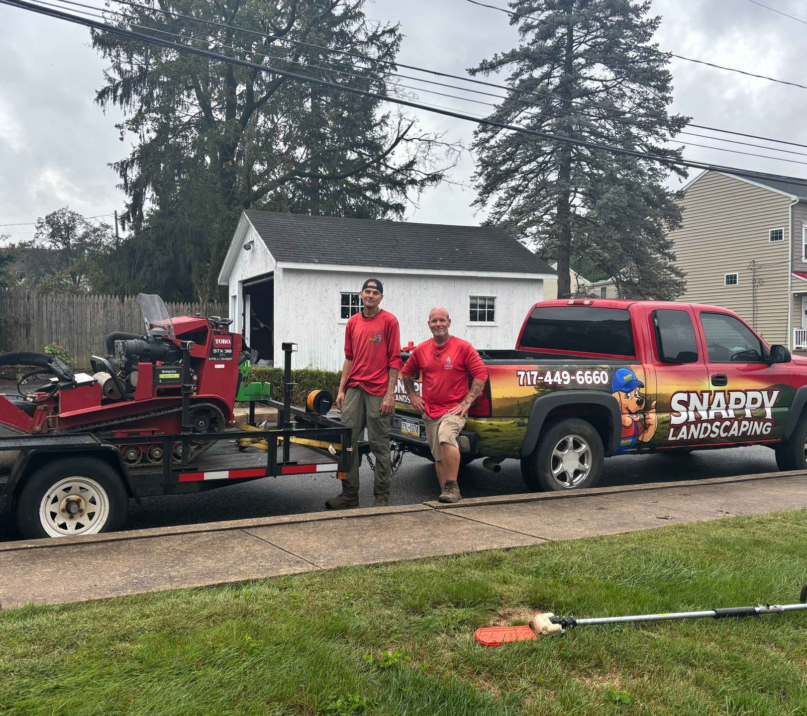 Two men in red shirts stand by a truck with a trailer holding a stump grinder.