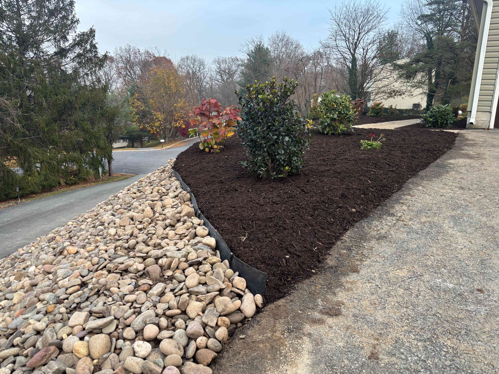 Landscaped driveway with river rocks, dark mulch, shrubs, and a gravel path beside a house