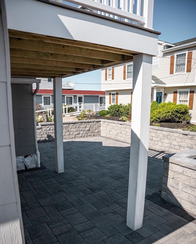 Covered patio with white posts and gray pavers beside stone retaining walls and apartment buildings