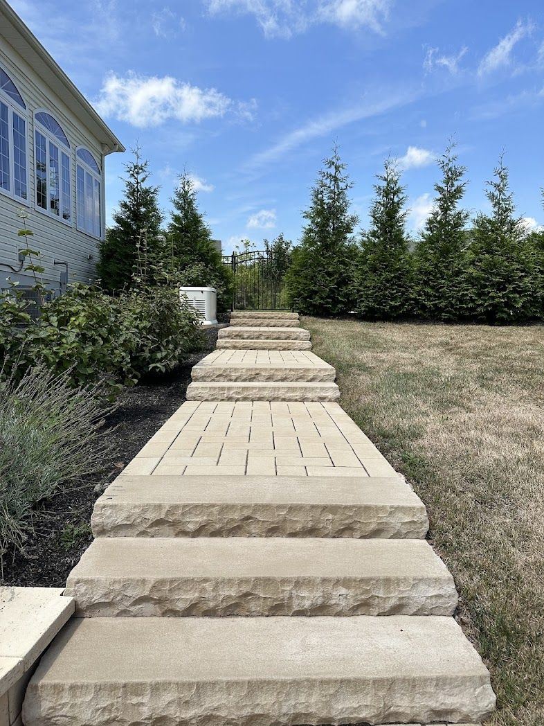 Stone walkway steps through a landscaped yard beside a house and evergreen trees under a blue sky.