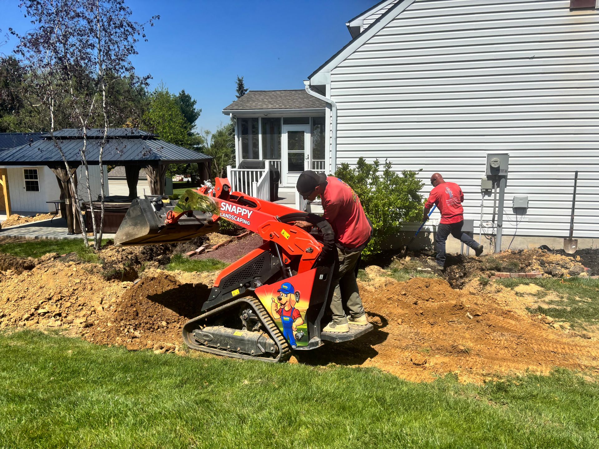 Outdoor Projects Workers using a red mini excavator to dig mulch in a yard beside a white house