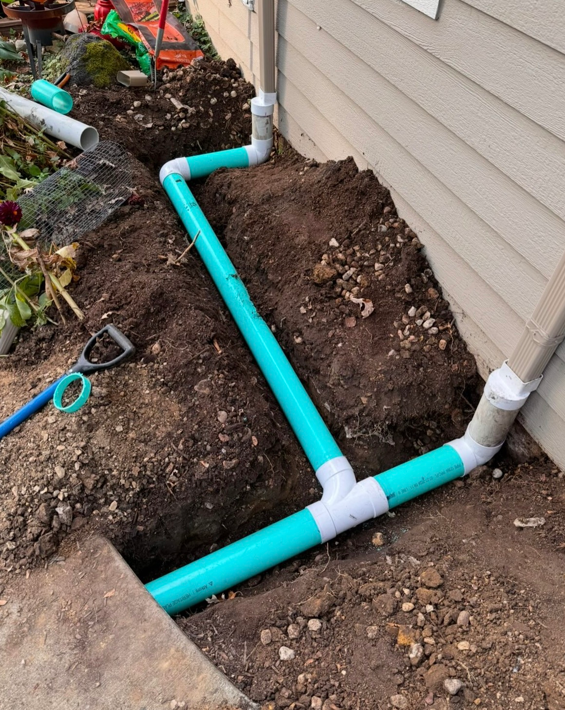 Bright turquoise PVC pipes laid in a dug trench along a house foundation, with white fittings and soil piled nearby