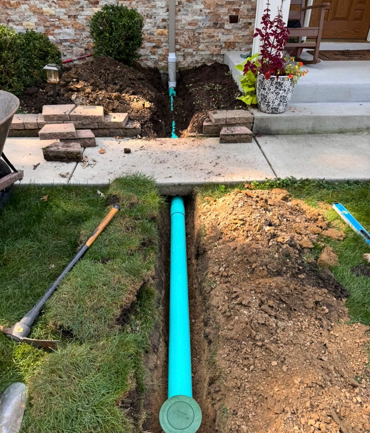 Trenched yard with bright blue pipe running under a sidewalk to a house entrance.