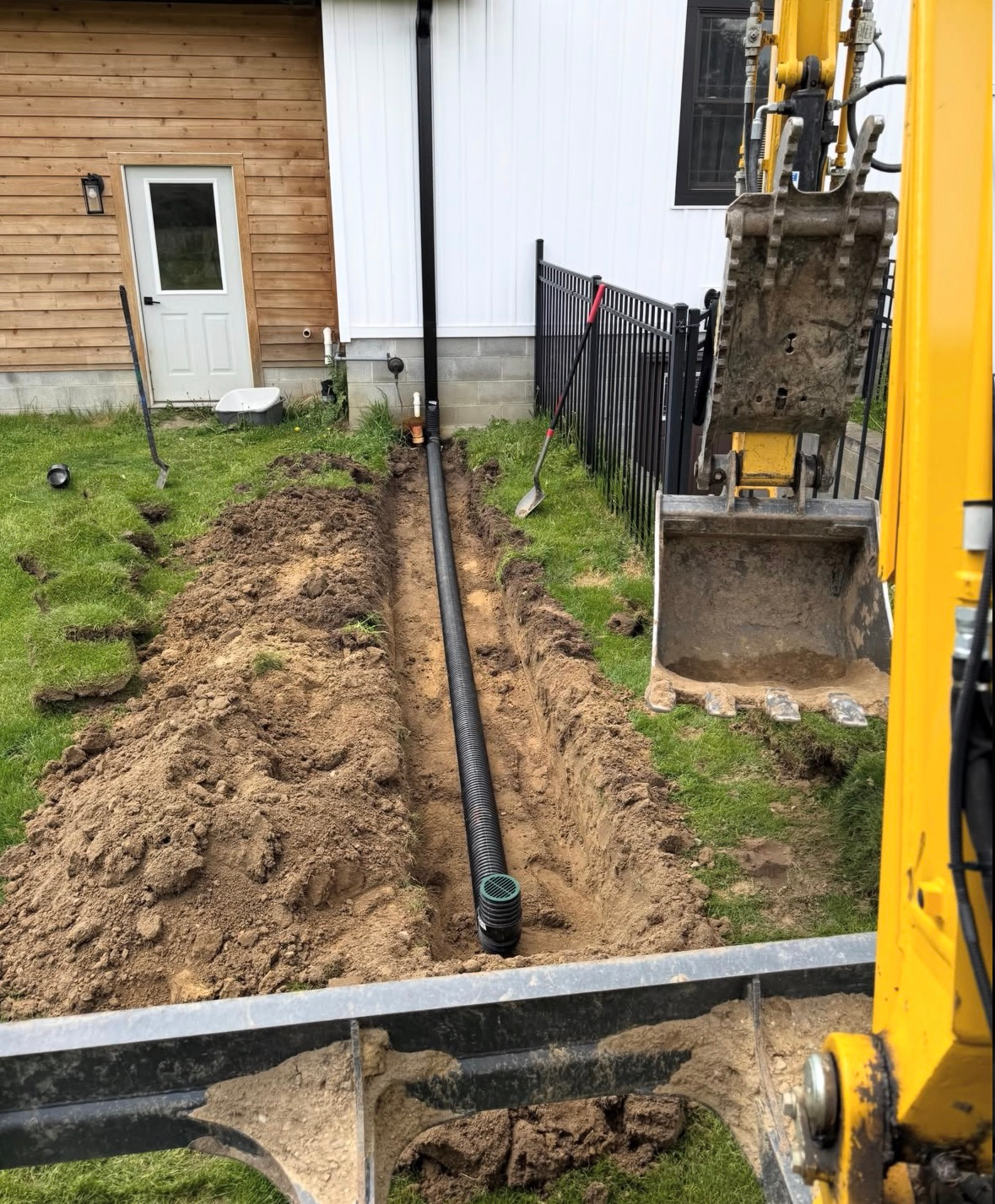 Trench beside a house with a black pipe laid in dirt, next to a backhoe and a white wall.