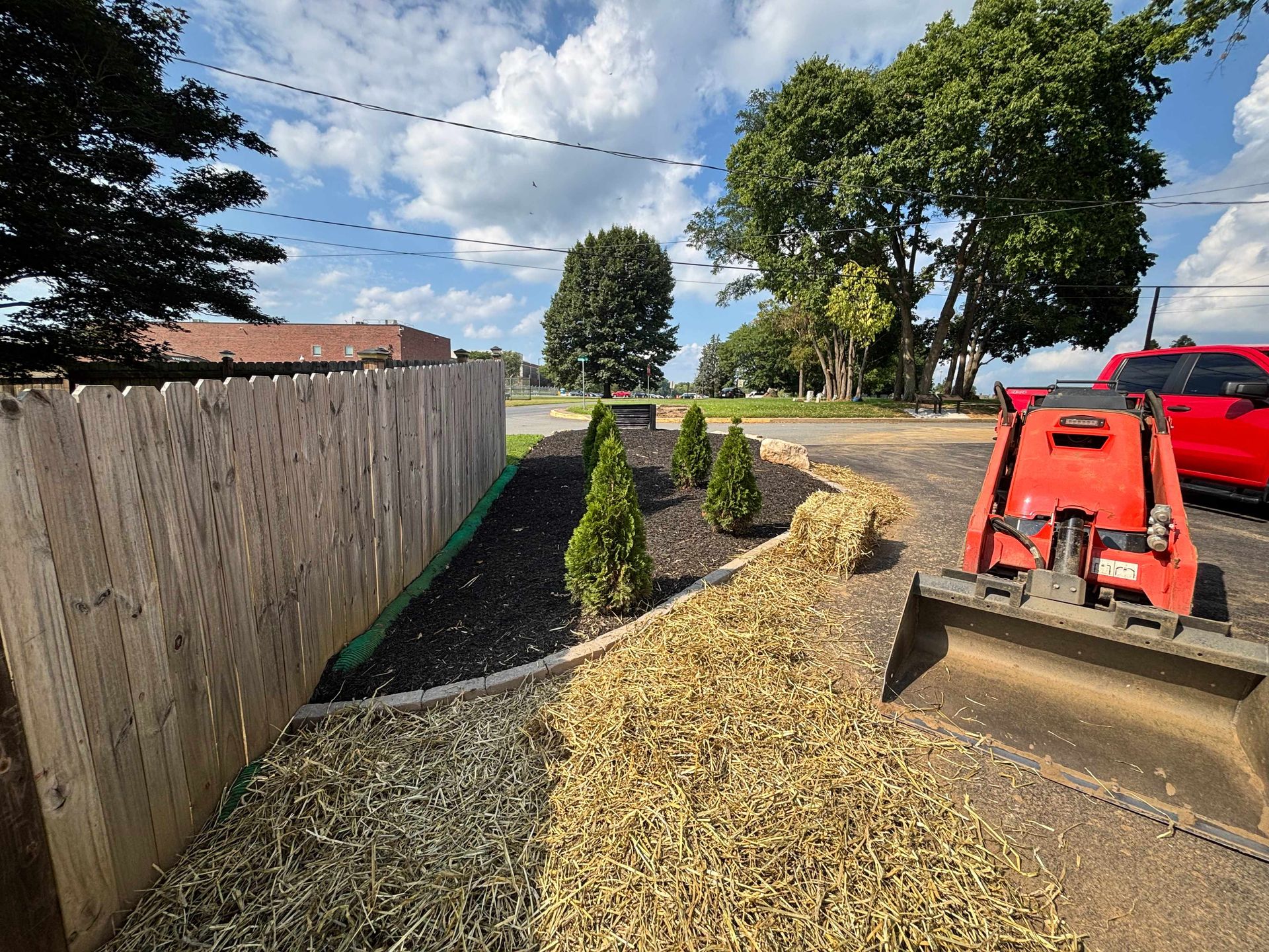 Freshly laid mulch beside a wooden fence, with small shrubs and a red tractor on a sunny residential lot