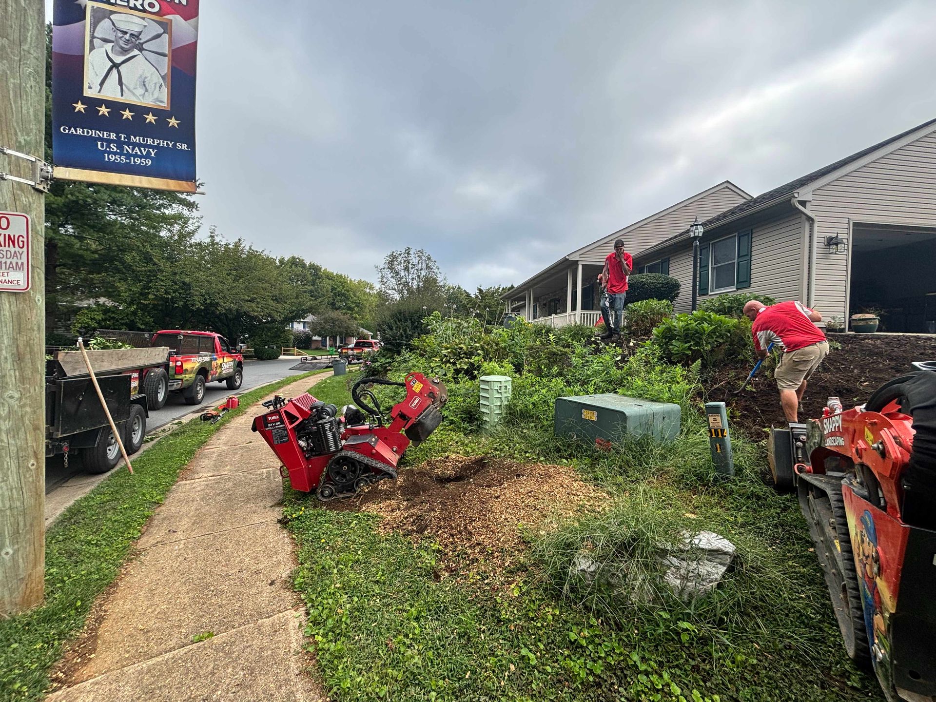 People clearing red debris along a path beside a building and greenery after a storm.
