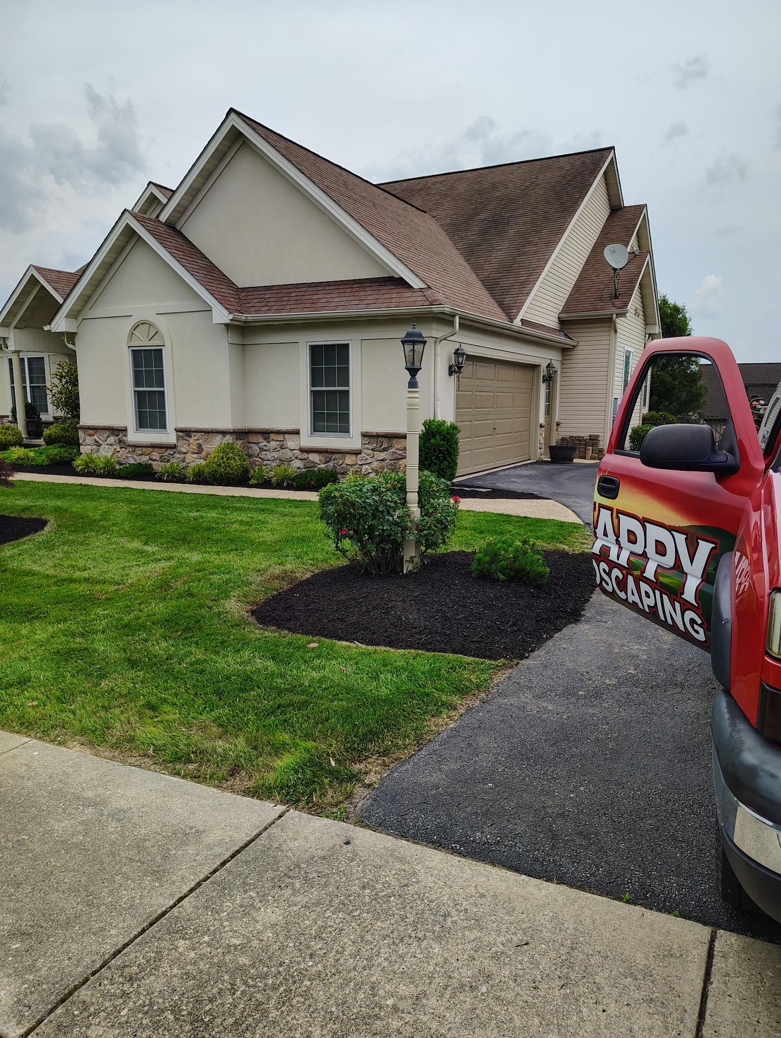 A red work truck parked on a driveway in front of a tan suburban house with brown roofing and landscaped mulch beds.