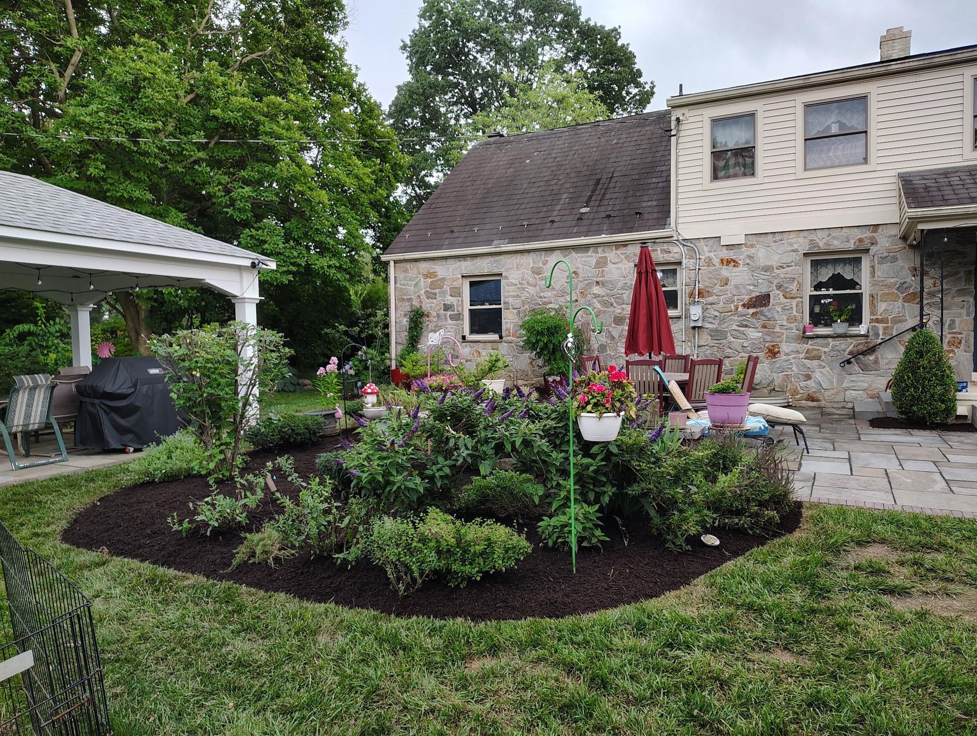 A landscaped stone house with a patio, a red umbrella, and a covered garden seating area in a backyard.