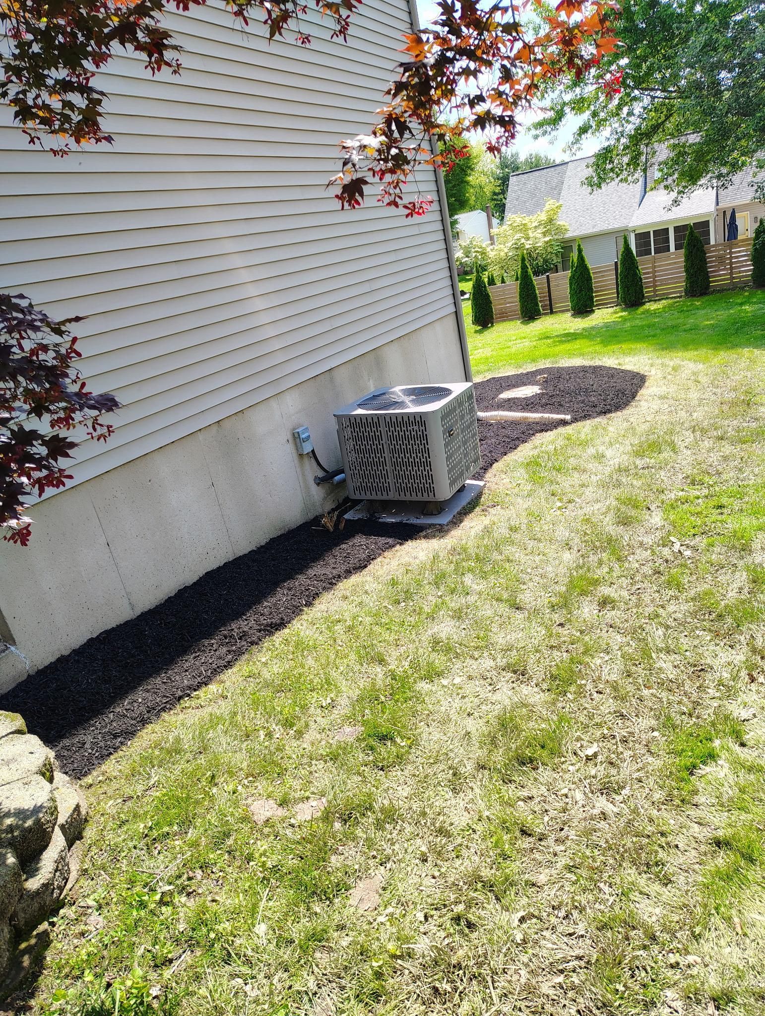 An outdoor air conditioning unit sits next to a light-colored house foundation surrounded by dark mulch and a grassy lawn.
