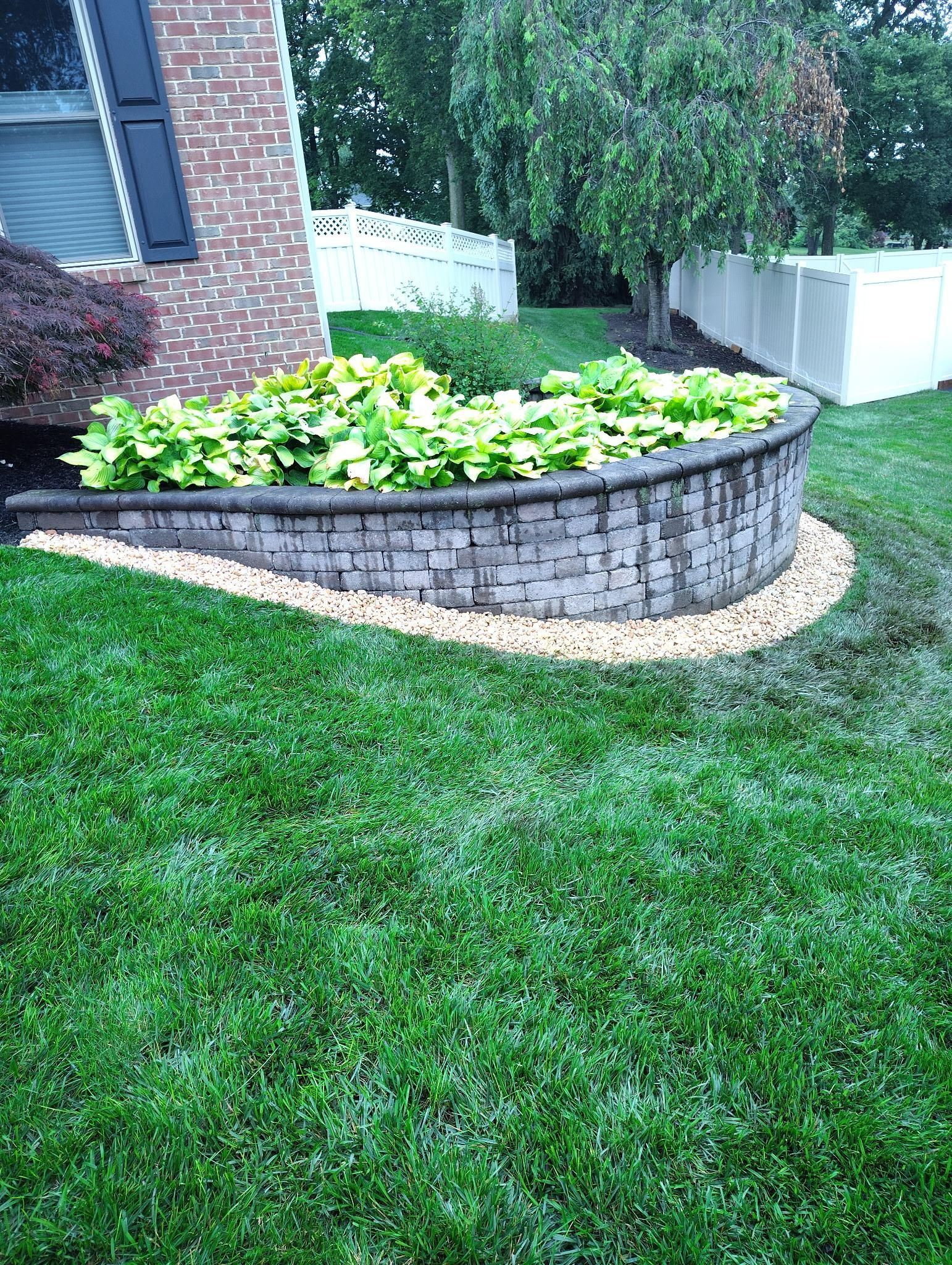 Curved stone planter bed with green plants beside a brick house and lawn