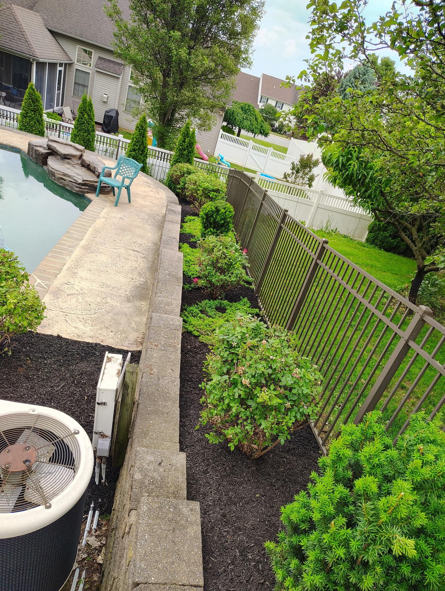 A landscaped backyard featuring a pool bordered by a stone wall, mulch beds with small shrubs, and a metal fence.