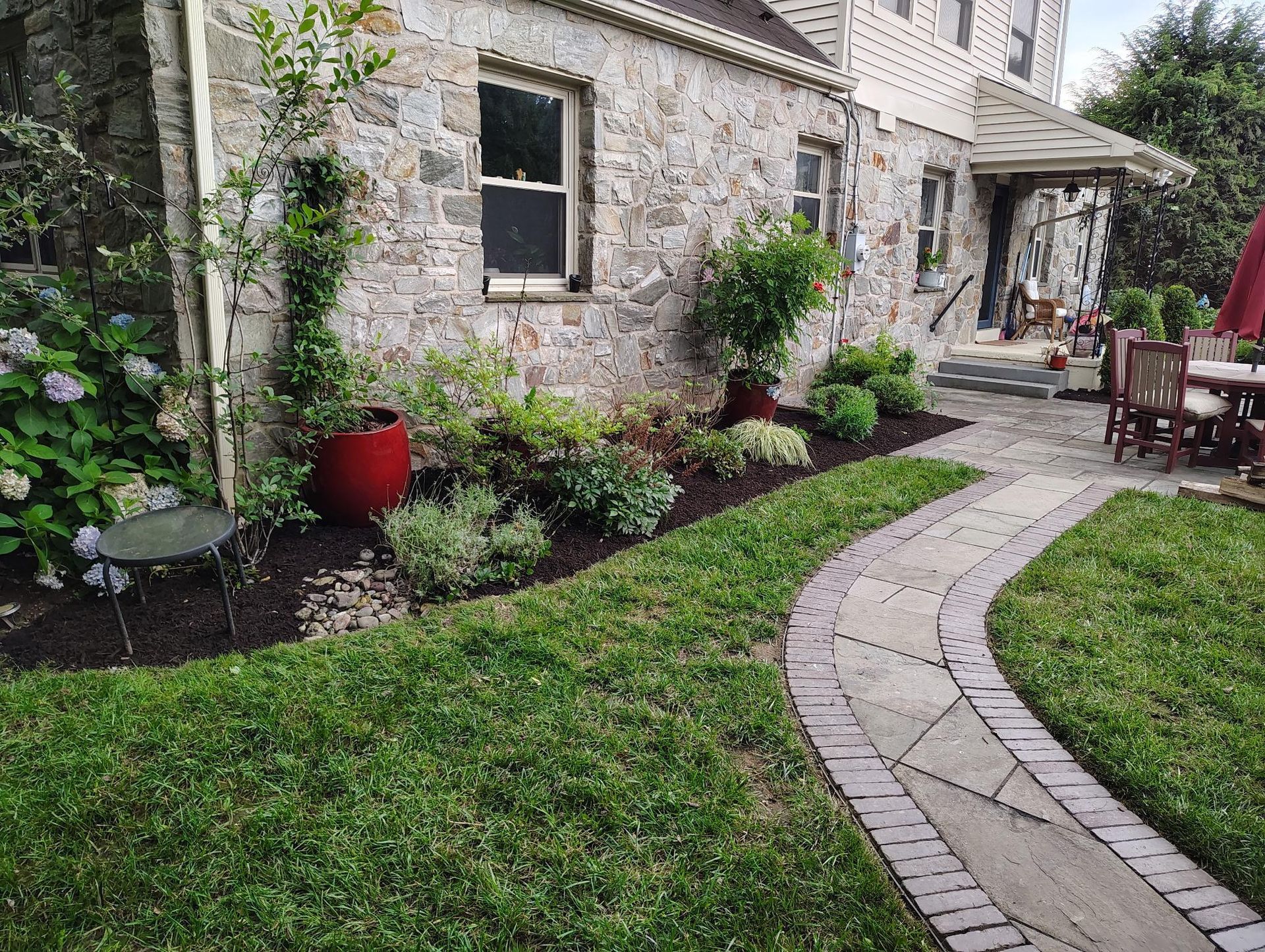 A stone building with a curved stone path leading to a patio, flanked by garden beds with shrubs and a red planter.