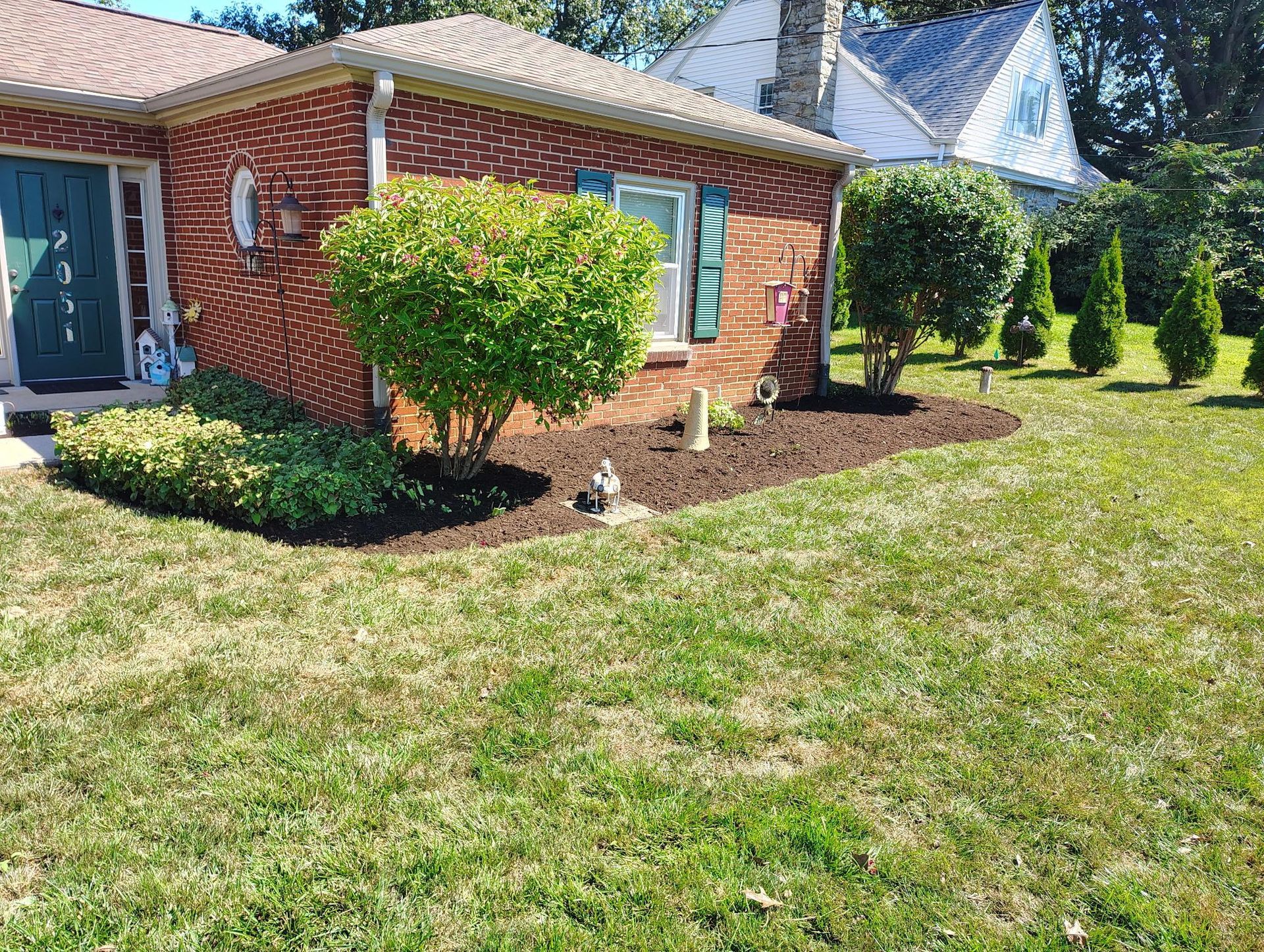 A brick house with a green door and shuttered window, featuring a neatly mulched garden bed and a sunny lawn.